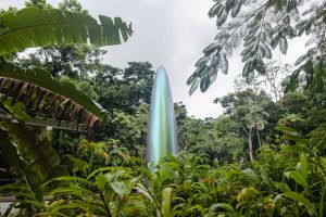 A monolithic green iridescent sculpture emerges from dense vegetation under a cloudy sky.