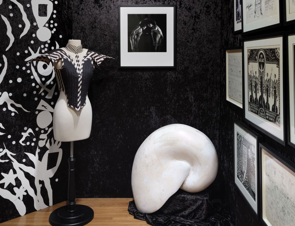 A small black-walled exhibition room displays comic-style drawings, a sculptural white shell form and a black-and-white striped garment mounted on a mannequin.