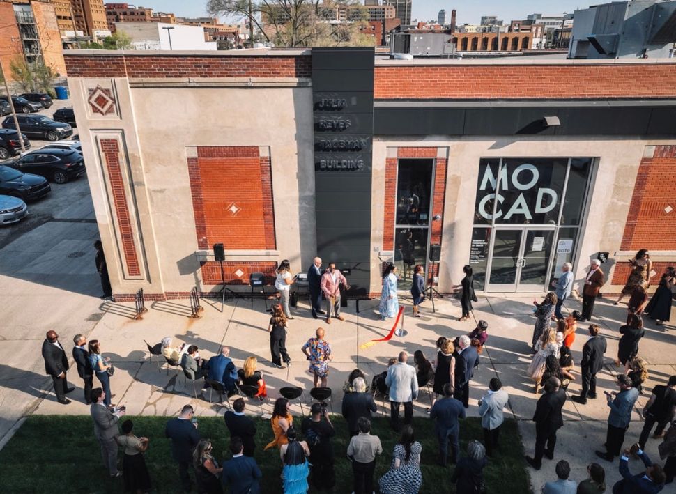 An overhead exterior photo shows a crowd gathered outside the MOCAD building for an event, with speakers, chairs and the museum’s entrance visible.