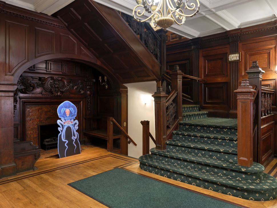 A glowing jellyfish-like sculpture is installed in front of a carved wooden fireplace beside a grand staircase inside the Estonian House.