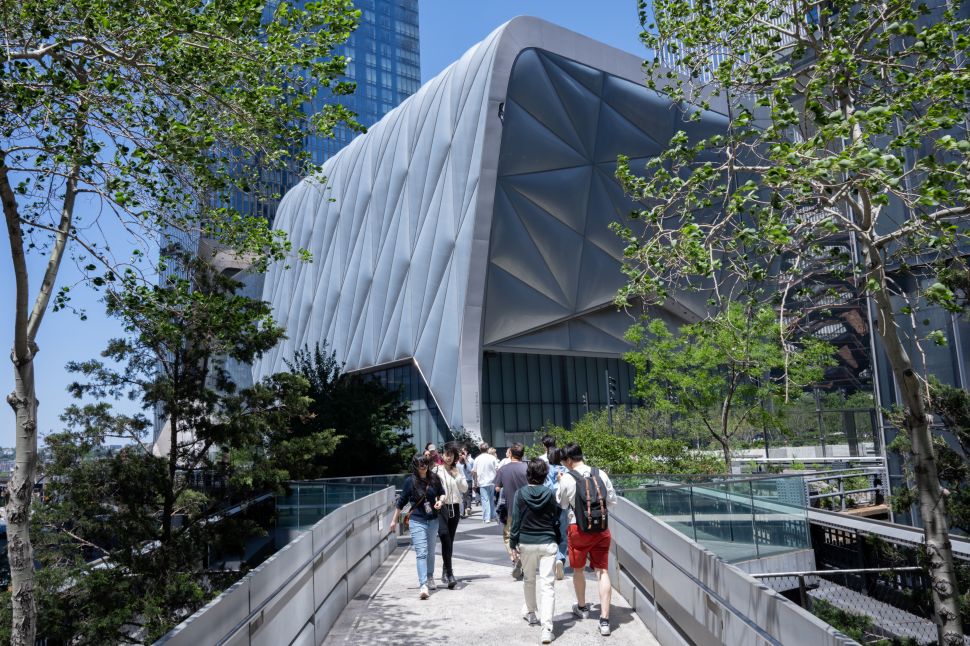 Visitors walk along an elevated outdoor path toward a large, modern, faceted building surrounded by trees and city structures.