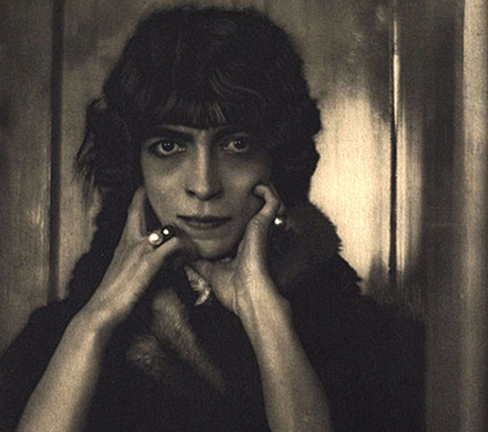 A close-up portrait of Marchesa Luisa Casati, her face framed by her hands, wearing a fur-trimmed garment and her signature heavily kohl-lined eyes. The sepia-toned photograph captures an unusual stillness — intense but unperformed, unlike nearly every other image she left behind.