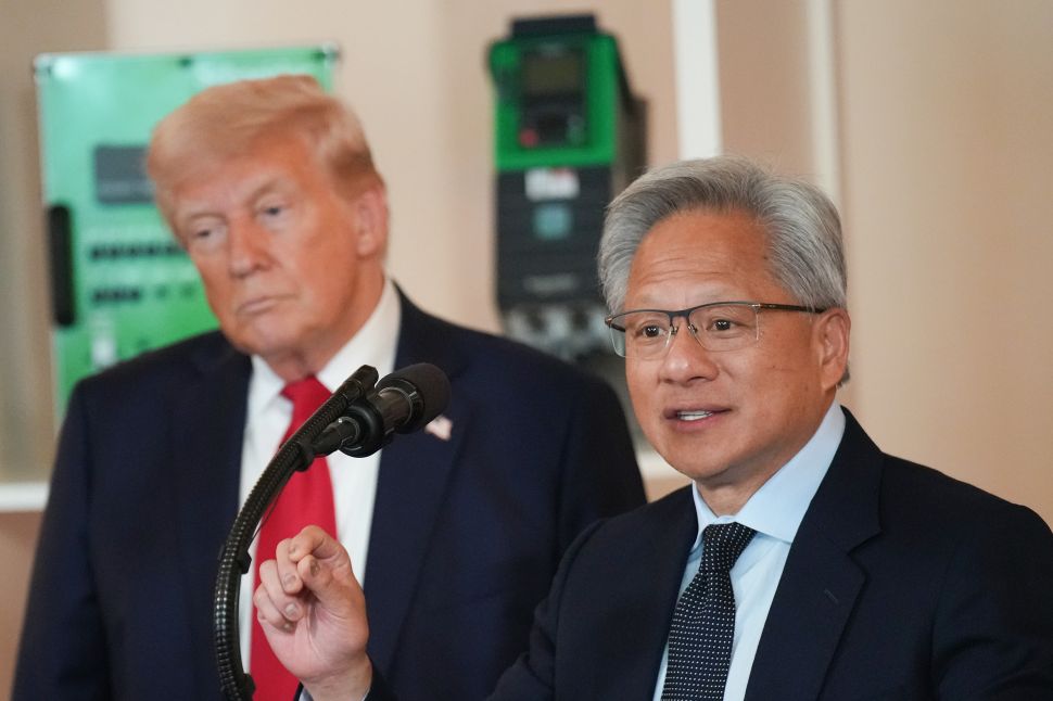 Man in suit and red tie stands behind another man in a suit and black tie speaking into a microphone 