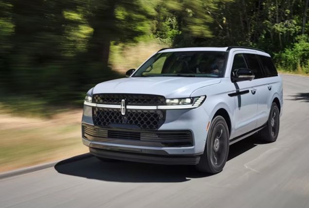 A white Lincoln Navigator SUV drives along a tree-lined road, with blurred greenery suggesting motion.