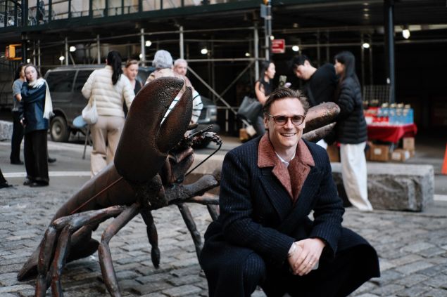 Man in glasses and black outfit crouches besides bronze lobster statue