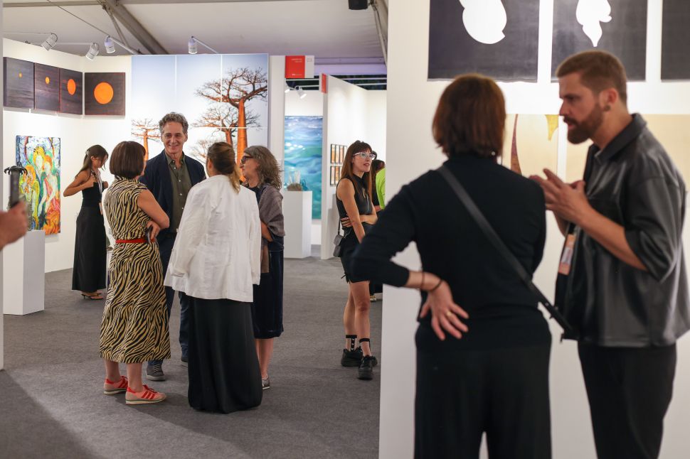 A group of visitors stands in a white-walled booth at the art fair, casually conversing while surrounded by colorful contemporary paintings and photography.