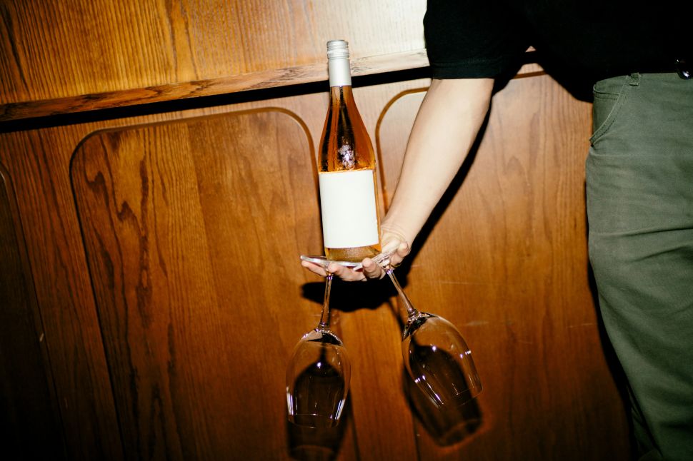 A wine bottle balances on a person's palm while two wine glasses hang upside down from their fingers