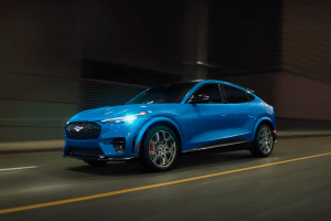 A blue Ford Mustang Mach-E drives through a city street at night beneath blurred urban lights.